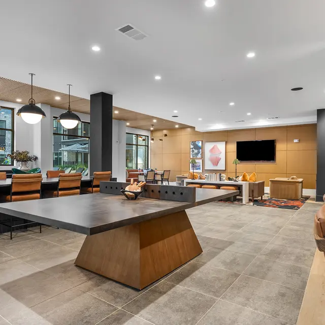 A modern lounge area featuring a large table, wooden and leather seating, pendant lights, and a wall-mounted TV.