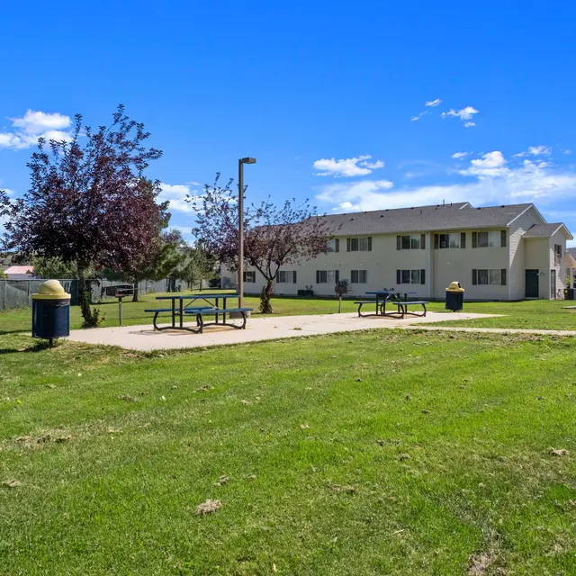 A spacious outdoor area featuring picnic tables, a playground, and green lawns, surrounded by trees and open spaces. The setting provides a perfect spot for residents to enjoy outdoor activities and relaxation.