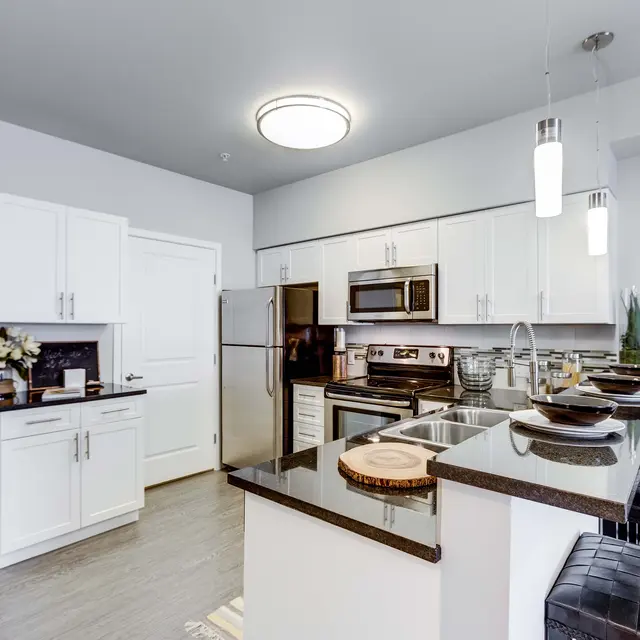 A modern kitchen with sleek white cabinetry, stainless steel appliances, and a black granite countertop. The kitchen features a small dining area with black bar stools and decorative plants.