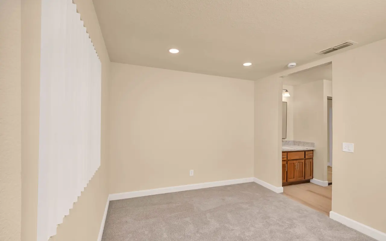 An empty room featuring light beige walls and carpet, with a window covered by white blinds. The room leads to a bathroom area with wooden cabinets visible.