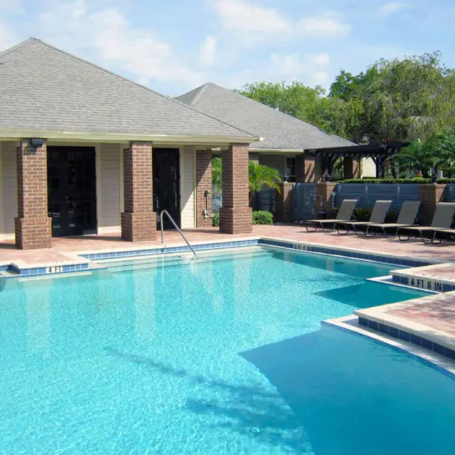 A clear blue swimming pool surrounded by lounge chairs and a well-maintained landscape, featuring a brick building in the background.