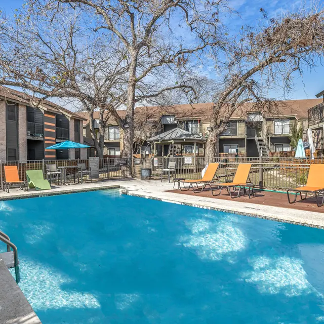 An outdoor gated swimming pool with lounge chairs near large trees and apartment buildings.