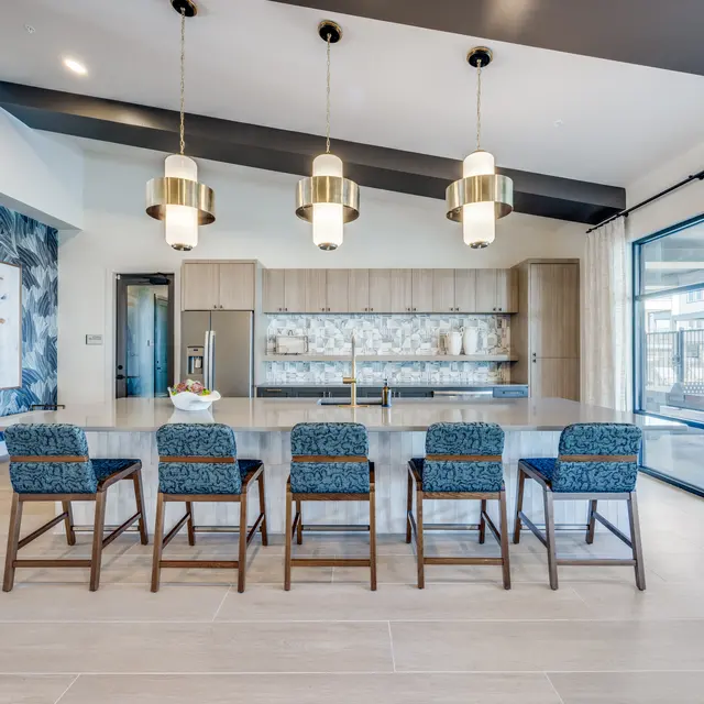 A modern kitchen featuring a long table with eight blue upholstered chairs, sleek cabinetry, and stylish pendant lights. Large windows allow for natural light and a view outside.