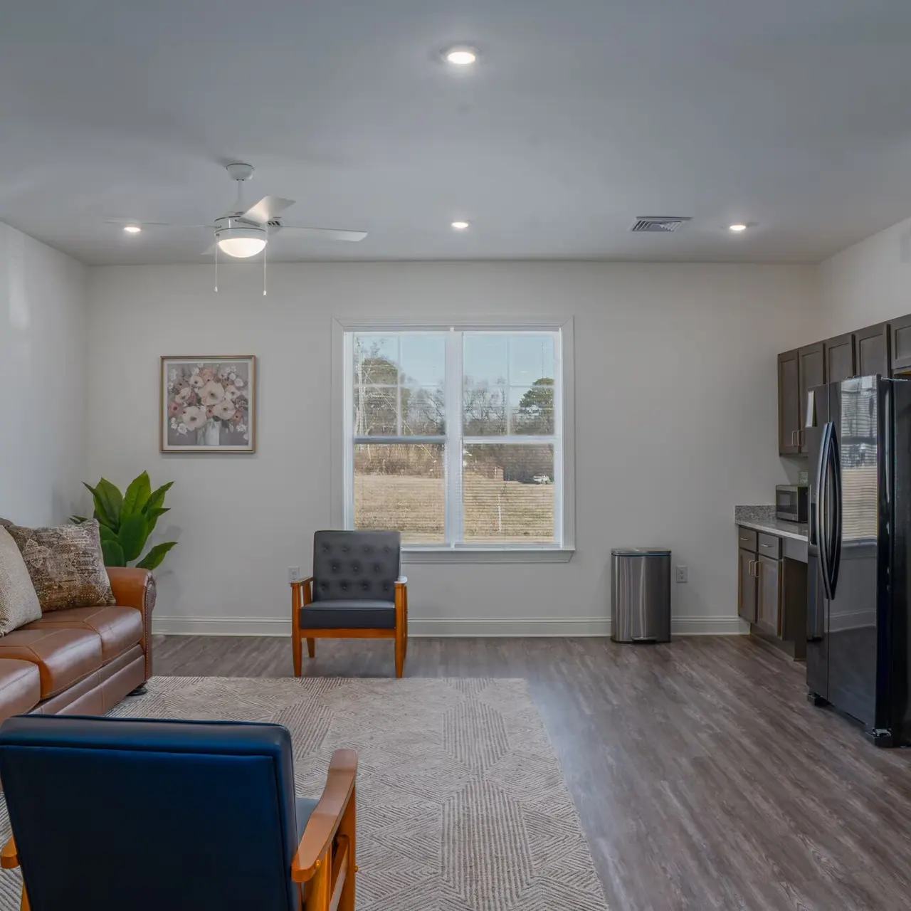 A bright, modern living room featuring a brown leather sofa with decorative pillows, a blue armchair, and a kitchen area with stainless steel appliances in the background. Large window allows natural light to illuminate the space.