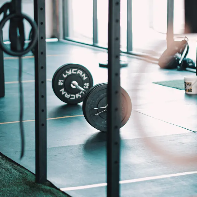 A gym scene featuring a barbell on the floor, framed by gym equipment and large windows in the background.