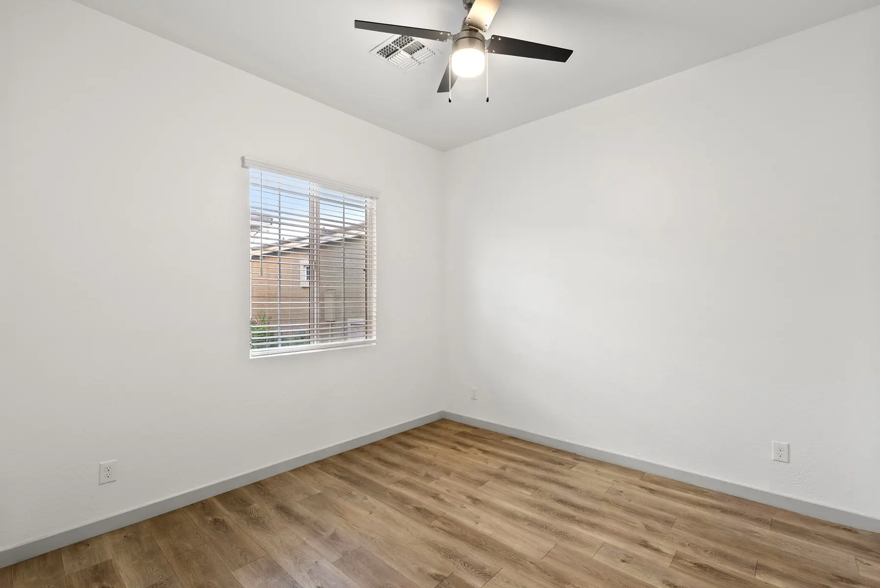 A small, empty room with a wooden floor, ceiling fan, and a window with blinds, featuring plain white walls.