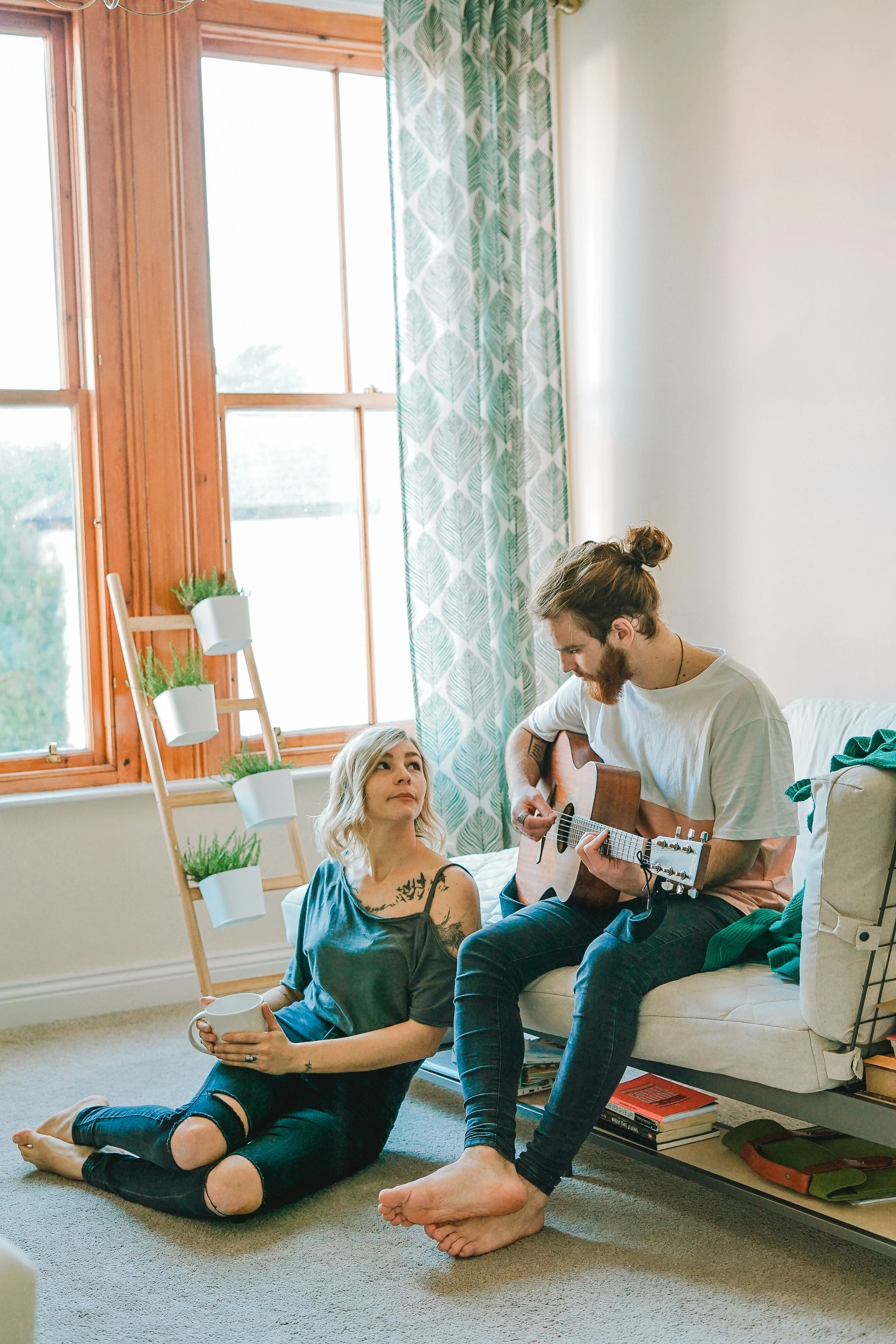 A couple in a cozy living room with a guitar. The man is sitting on a couch playing the guitar while the woman sits on the floor, looking up at him. Natural light filters in through large windows.
