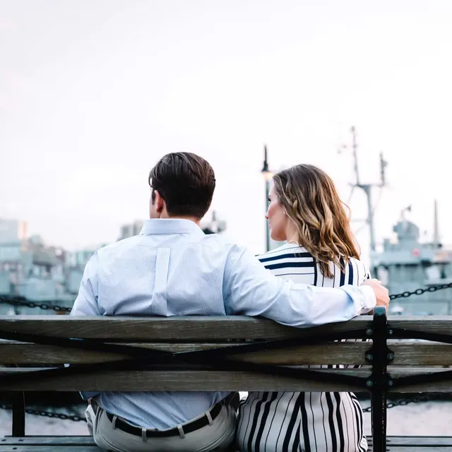 A couple sitting on a bench, facing away, with ships in the background, enjoying a scenic view together.