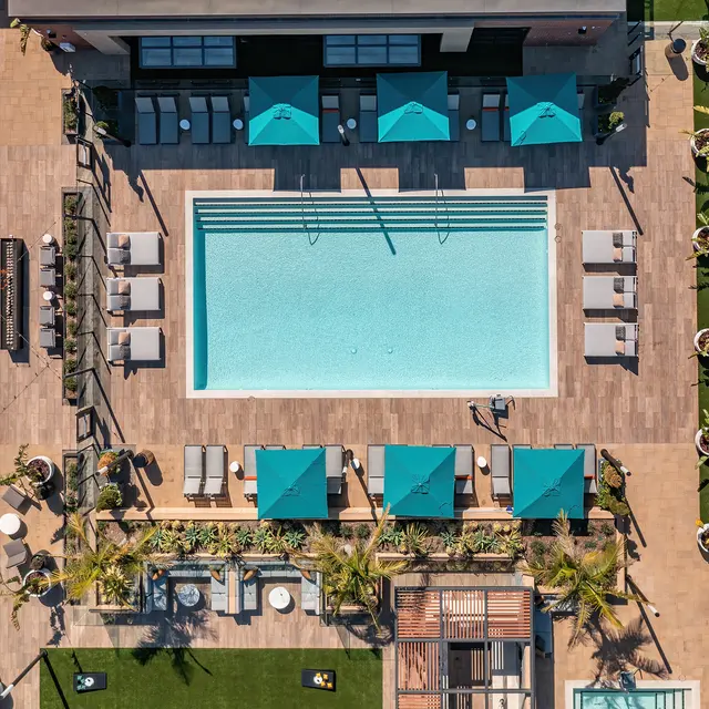 Aerial view of a modern pool area featuring a rectangular swimming pool surrounded by lounge chairs and cabanas, with turquoise umbrellas providing shade, and landscaped greenery.