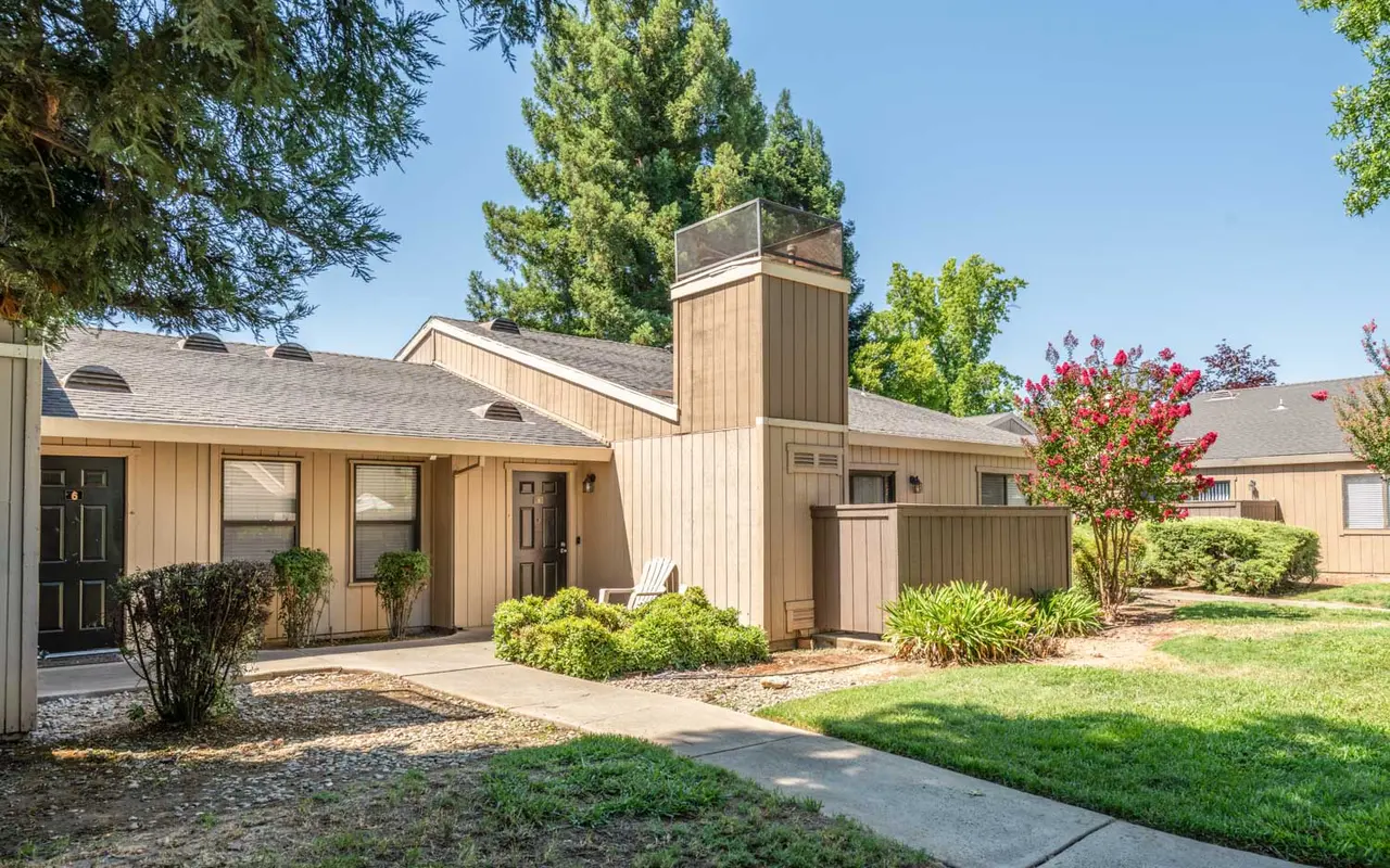 A view of a residential townhouse complex with beige wooden siding, featuring several units on either side of a concrete pathway. There are green bushes and trees in the foreground, and the sky is clear and blue.