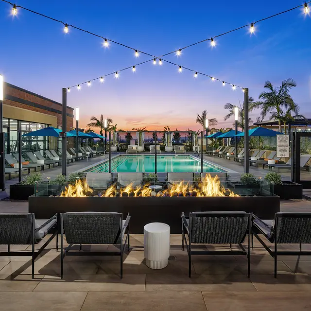 A beautifully lit rooftop pool area at sunset, featuring lounge chairs, fire pits, and palm trees.