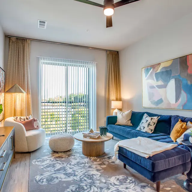 A modern living room featuring a blue sofa, a round coffee table, and a cozy chair. There are plants and a painting on the wall, with large windows allowing natural light to brighten the space.