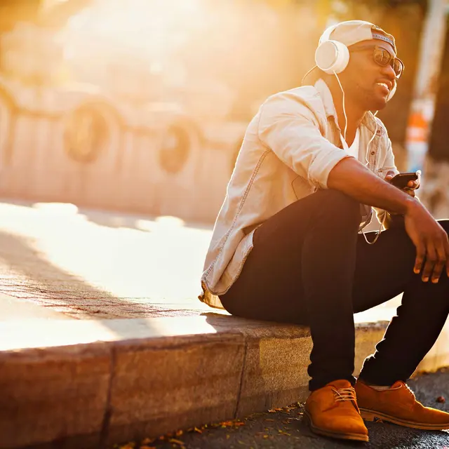 Chilling Outdoors with Music A young man sitting outdoors, enjoying music through headphones while basking in the sunlight. He is dressed casually in a light shirt and dark pants.