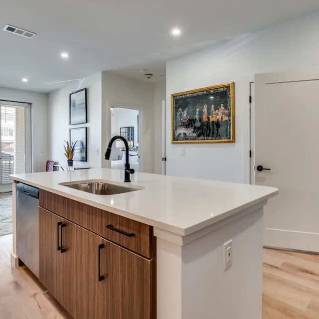 A modern kitchen with an island, featuring a sink and wooden cabinetry, opening into a living area with hardwood floors and large windows. There's a framed artwork on the wall and a laundry area visible in the background.