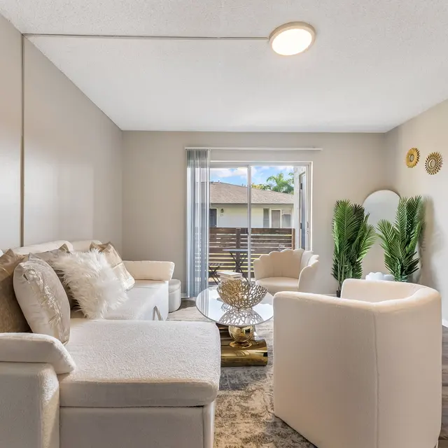A stylish modern living room featuring a white sectional sofa and two white armchairs. Decorative pillows are on the sofa. A coffee table with a decorative bowl sits in the center. There are green plants and wall decor with circular mirrors. Large windows provide natural light.