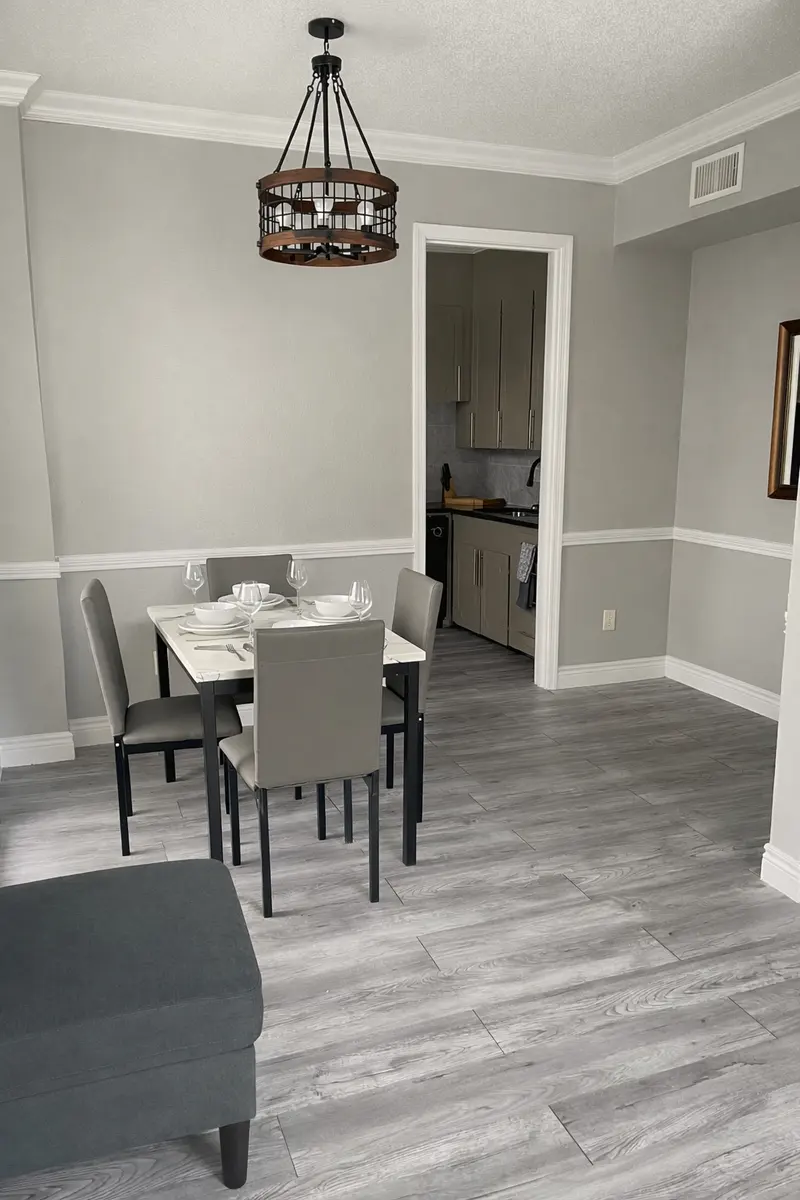 A stylish dining area featuring a wooden table with four chairs, next to a gray couch. A pendant light hangs above the table, and a doorway with kitchen appliances is visible in the background.