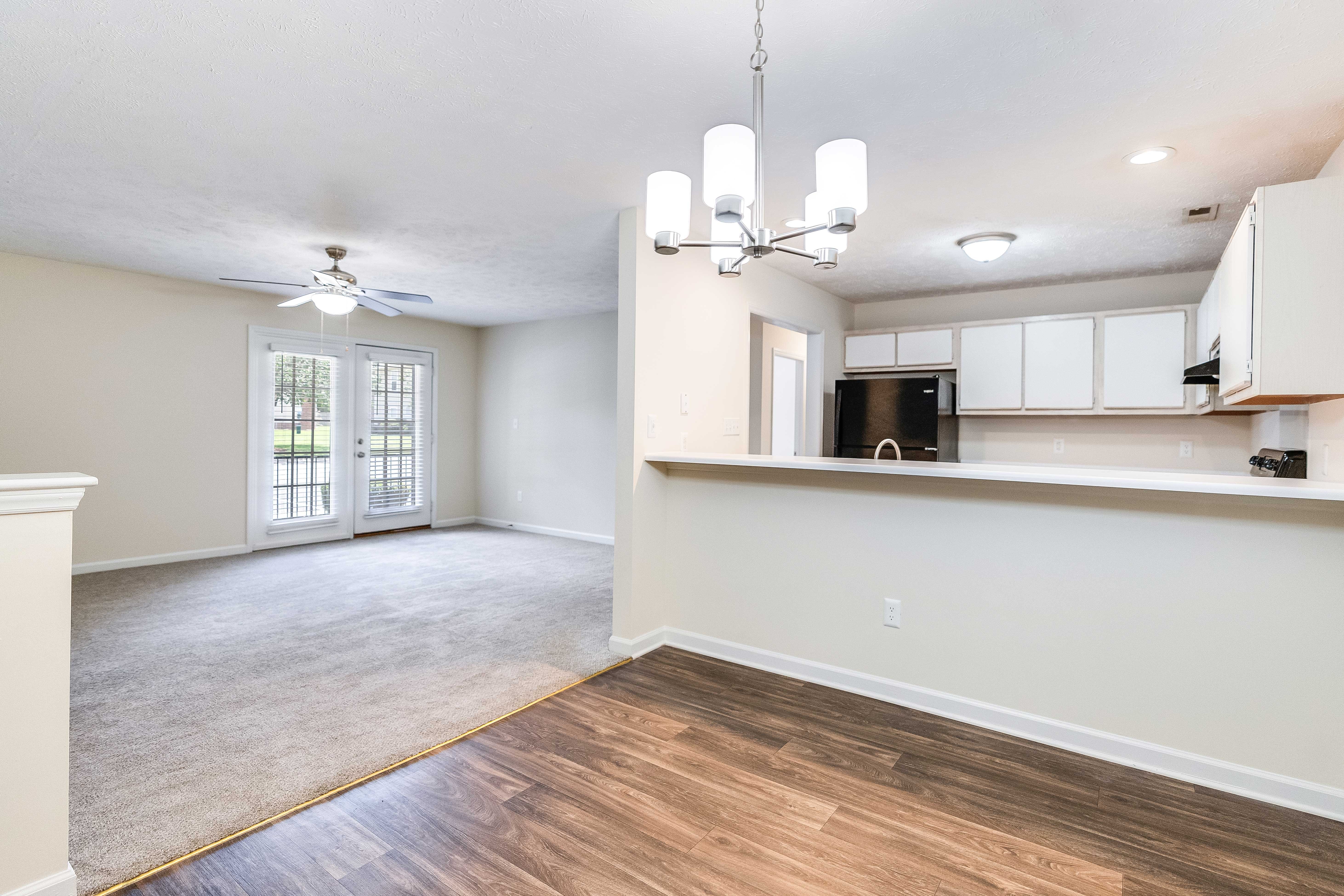 A spacious living area with a light-colored carpet, a ceiling fan, and large windows leading to a patio. The adjoining kitchen features white cabinets and a breakfast bar, with a black refrigerator and microwave visible.
