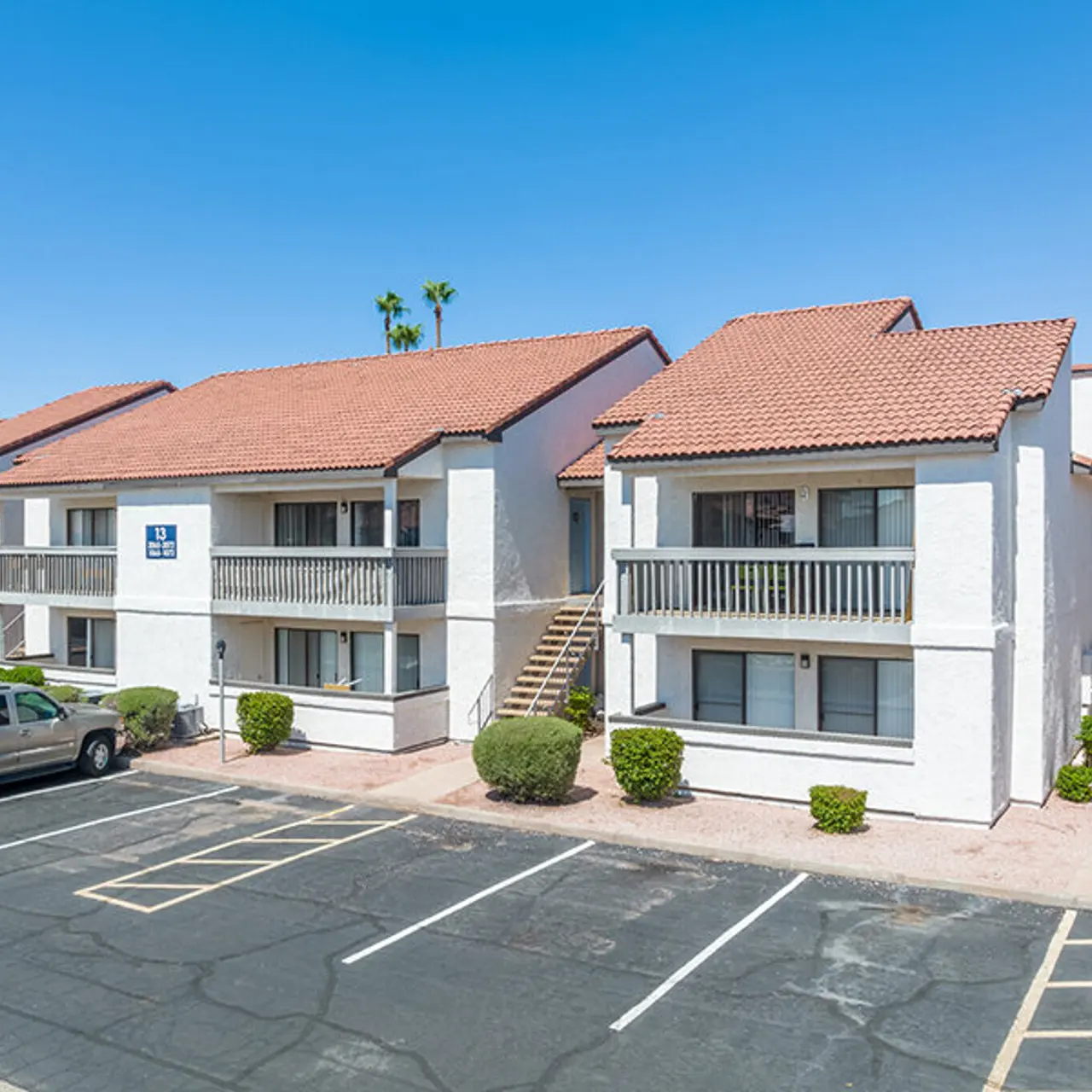 Exterior view of a two-story apartment complex with a parking lot, featuring a white stucco facade and a tile roof, surrounded by small shrubs and palm trees under a clear blue sky.