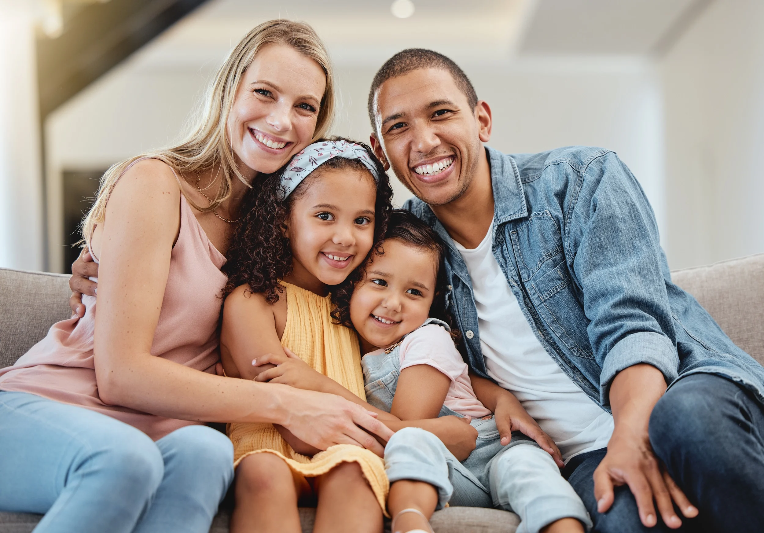 A joyful family of four consisting of two adults and two young children sitting closely together on a sofa, all smiling and posing for the photo.