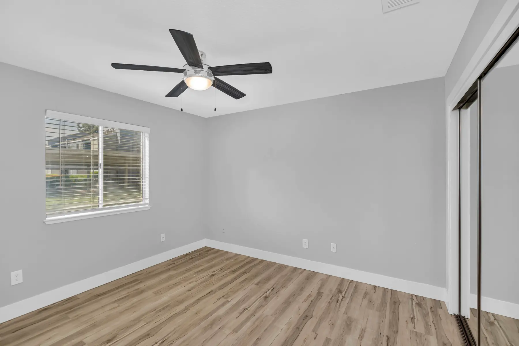 A modern bedroom featuring light gray walls, a ceiling fan with five blades, and a large window with blinds. The floor is made of wood, and there is a mirrored closet door visible on the right.