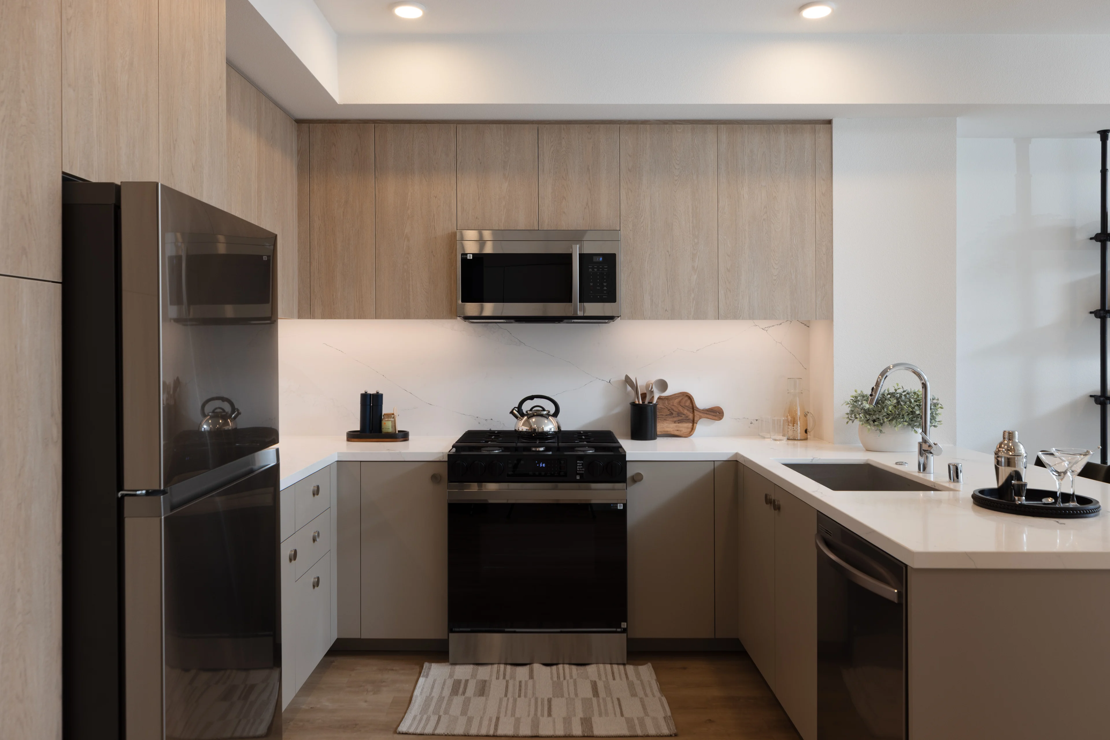 Elegant Modern Kitchen A modern kitchen featuring sleek cabinetry, a stainless steel refrigerator, a gas stove, and a microwave. The countertop is white with a subtle pattern, and the floor is light wood. A small decorative rug is placed in front of the stove, and there are plants and kitchenware visible on the countertop.