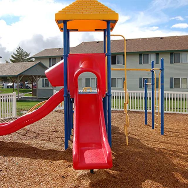 Children's Playground with Slide A colorful playground featuring a red slide, climbing structure, and safety fence, surrounded by grass and buildings in the background under a blue sky.