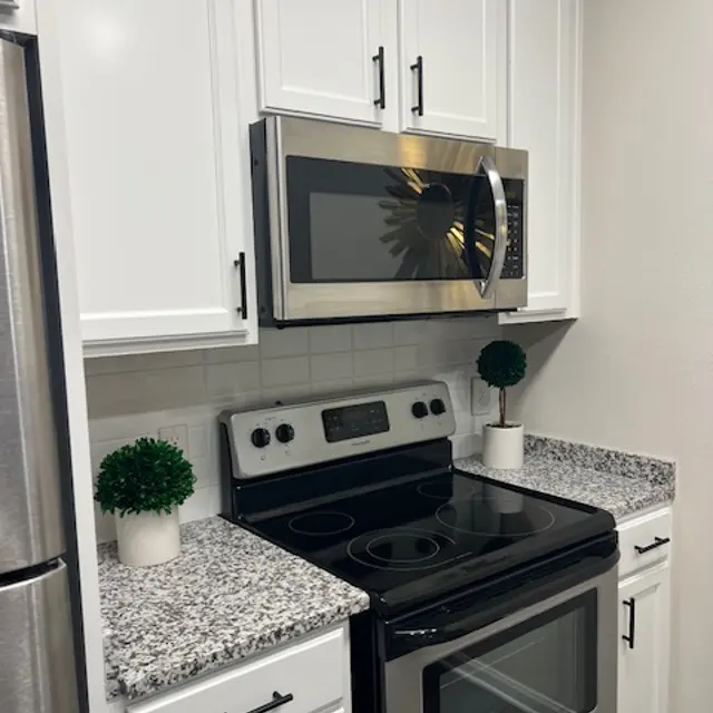 A modern kitchen with white cabinets, granite countertops, a black oven and stovetop, a metallic microwave, and decorative plants.