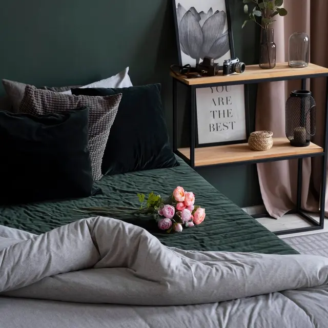 A cozy bedroom scene featuring a bed with gray bedding and dark green pillows. A bouquet of pink roses and greenery is placed on the bed. Behind, there is a small wooden shelf with decorative items, including a framed quote that says 'YOU ARE THE BEST.' The wall is painted dark green, and soft curtains are draped beside a window.