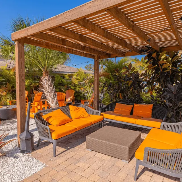 A cozy outdoor lounge area with orange cushions under a wooden pergola, surrounded by palm trees and lush greenery.