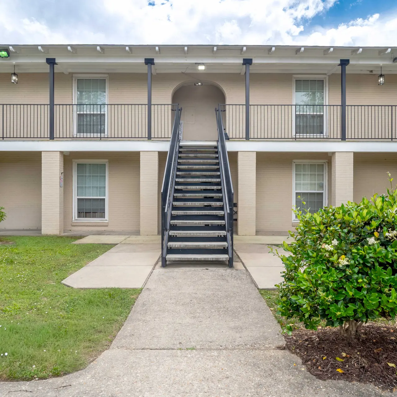 View of an apartment complex entrance featuring a staircase, greenery, and multiple windows.
