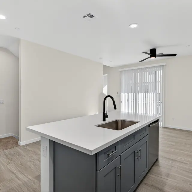 A modern apartment interior featuring a central kitchen island with a sink, grey cabinetry, and light-colored walls. The space is bright with natural light and has a ceiling fan and sliding glass doors looking out to a patio area.