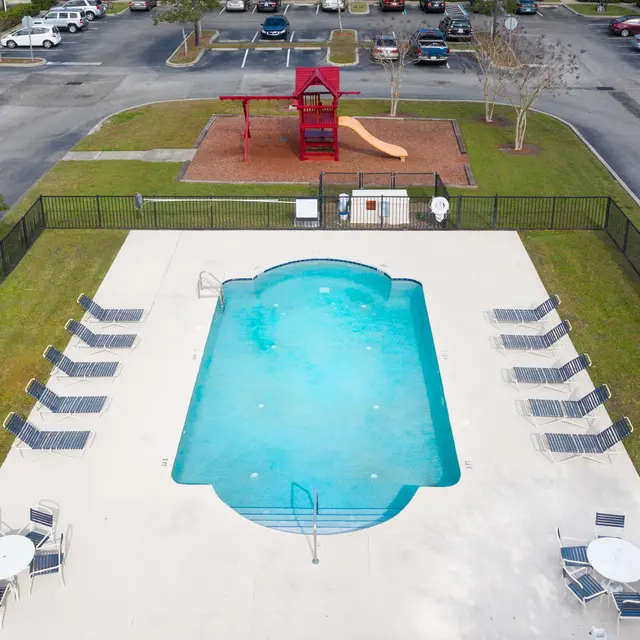 Aerial view of a community pool with a fenced area, lounge chairs, and adjacent playground equipment including a slide.