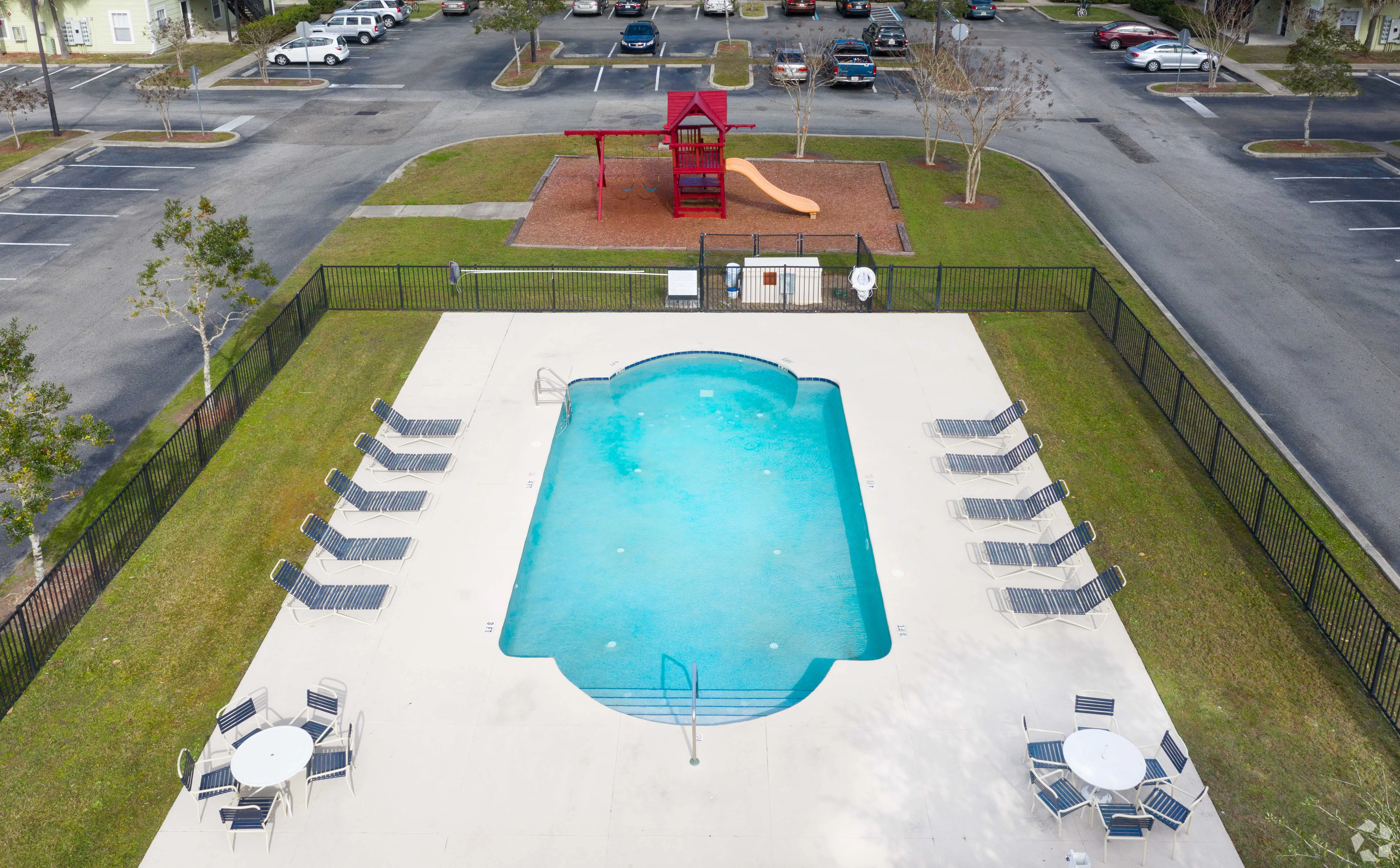 Aerial view of a community pool with a fenced area, lounge chairs, and adjacent playground equipment including a slide.