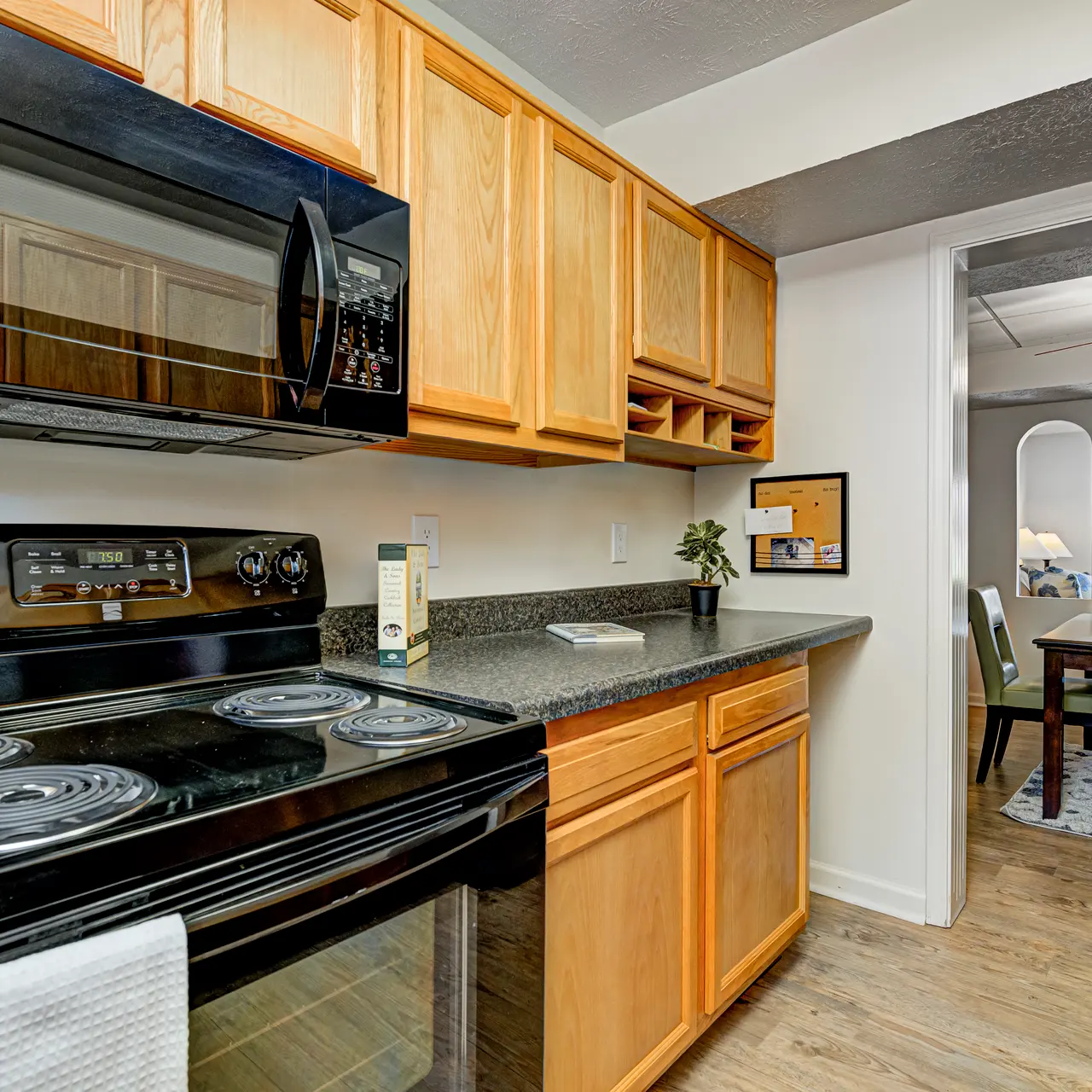 Modern Kitchen Interior A modern kitchen featuring wooden cabinets, a dark countertop, and black appliances including a microwave and stove. The kitchen opens to a dining area with a ceiling fan, and a glimpse of a dining table with floral decor can be seen in the background.