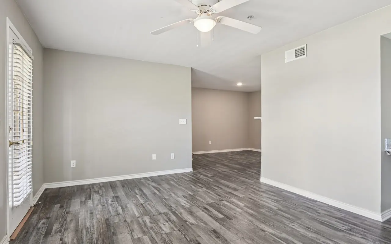 A spacious and bright living room with gray walls and wooden flooring. It features a ceiling fan and natural light coming from a door on the left side.
