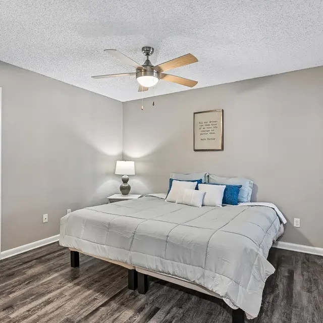 Modern Bedroom Interior A modern bedroom featuring a light gray bedspread on a bed with a wooden frame, a nightstand with a lamp, and a decorative wall art. The walls are painted in a neutral shade and there is a ceiling fan. Natural light filters through the window with blinds.
