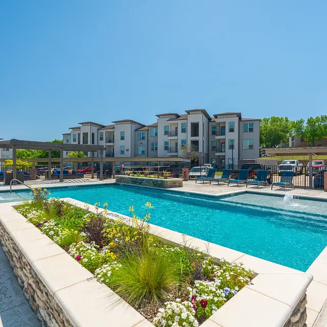 A sunny residential pool area with a swimming pool, surrounded by apartments and green landscaping.