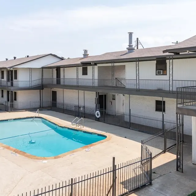 Apartment Complex with Pool An apartment complex featuring multiple two-story buildings surrounding a swimming pool. The pool is fenced and has lounge space around it, while the buildings are painted in light colors with balconies and air conditioning units visible. The scene is set on a sunny day.