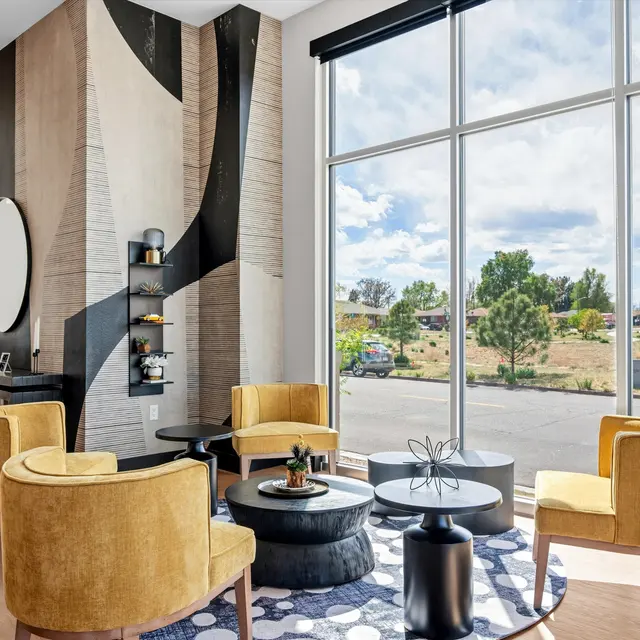 A stylish lounge area with large windows, featuring four yellow armchairs surrounding a circular coffee table. The walls have an artistic design, and a mirror is mounted on the wall. The view outside includes greenery and a clear sky.