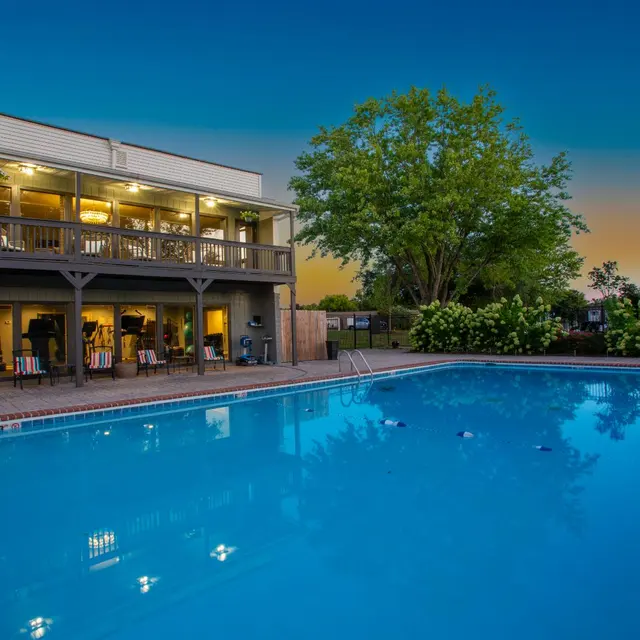 Evening Poolside View A swimming pool with lights reflecting in the water, surrounded by a building and greenery at dusk.
