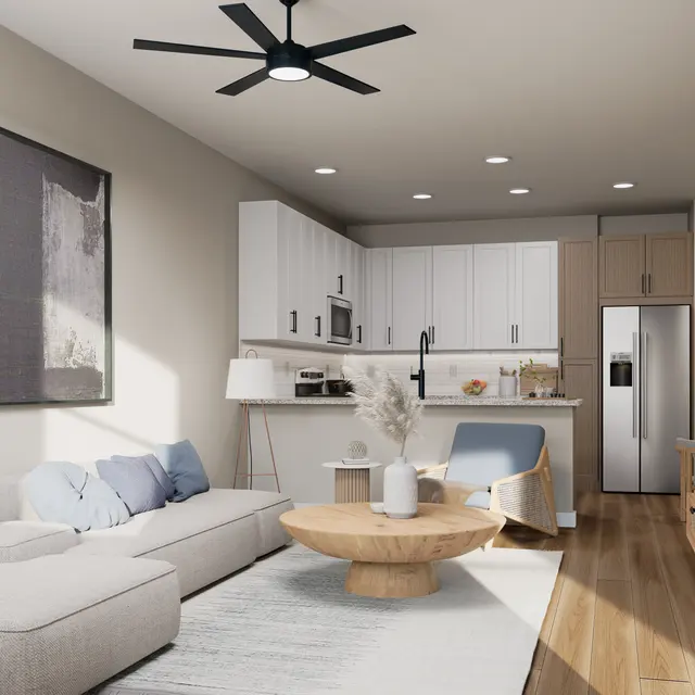 A modern living room featuring a light-colored sofa with pillows, a circular wooden coffee table, and a stylish ceiling fan. The room has a contemporary kitchen area adjoining it, with white and wood cabinetry, and stainless steel appliances. Natural light floods the space, enhancing the warm wood flooring and minimalist decor.