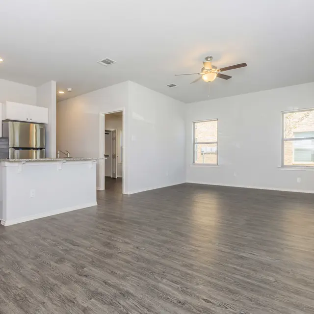 Spacious living area with open kitchen and large windows allowing natural light to enter, featuring light-colored walls and wood-like flooring.