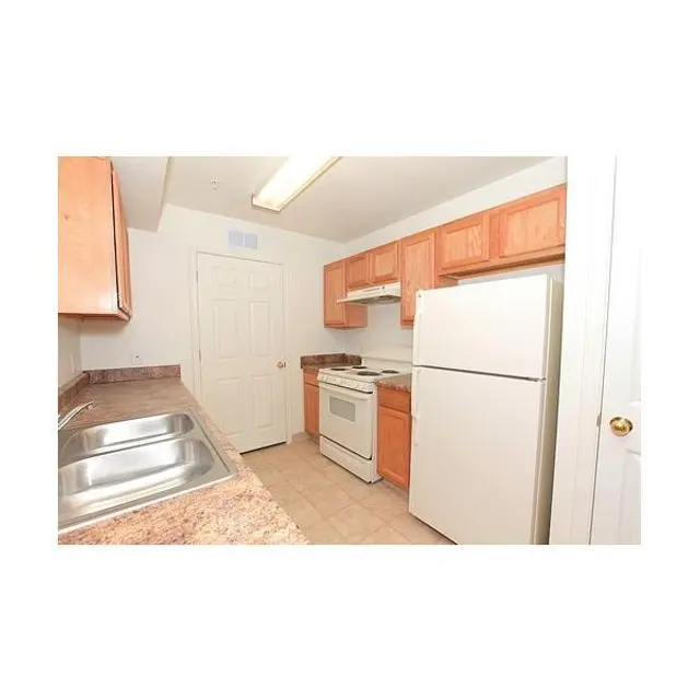 Compact Kitchen Interior A compact kitchen featuring light wooden cabinetry, a white refrigerator, a white stove, a double sink with a granite countertop, and a door leading to another room.
