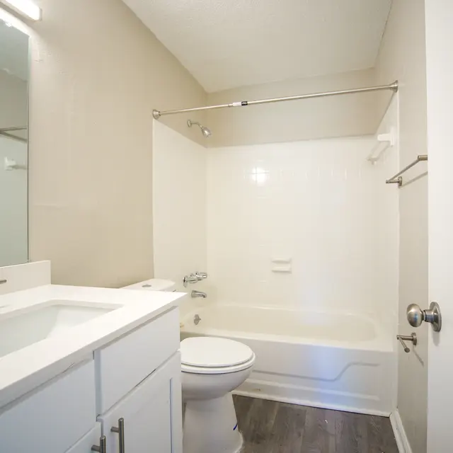 A modern bathroom featuring a white vanity with a sleek sink, a toilet, and a shower-tub combination enclosed by a white shower curtain. The walls are painted a soft beige, and the flooring is dark wood-like laminate. A mirror is mounted above the sink, and there is a bright light fixture above it.