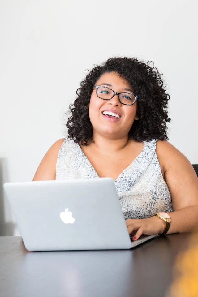 Smiling Woman Working on Laptop A smiling woman with curly hair and glasses sitting at a desk with a laptop. She wears a light-colored dress and appears happy and engaged.