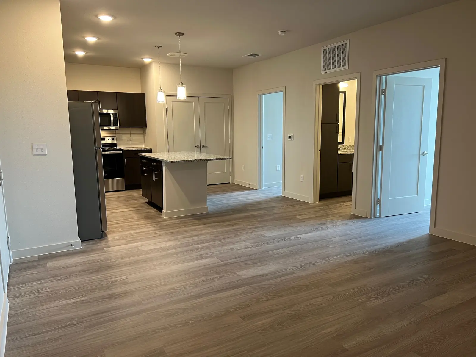 A spacious, modern apartment interior featuring an open layout with hardwood flooring, light-colored walls, and kitchen appliances visible in the background.