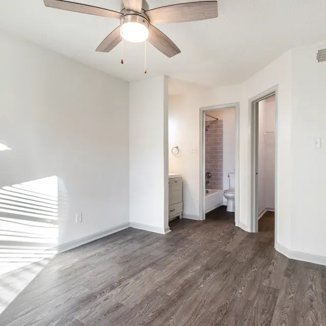 A spacious, empty room with a ceiling fan, wooden flooring, and light streaming through the window, leading to an adjoining bathroom.