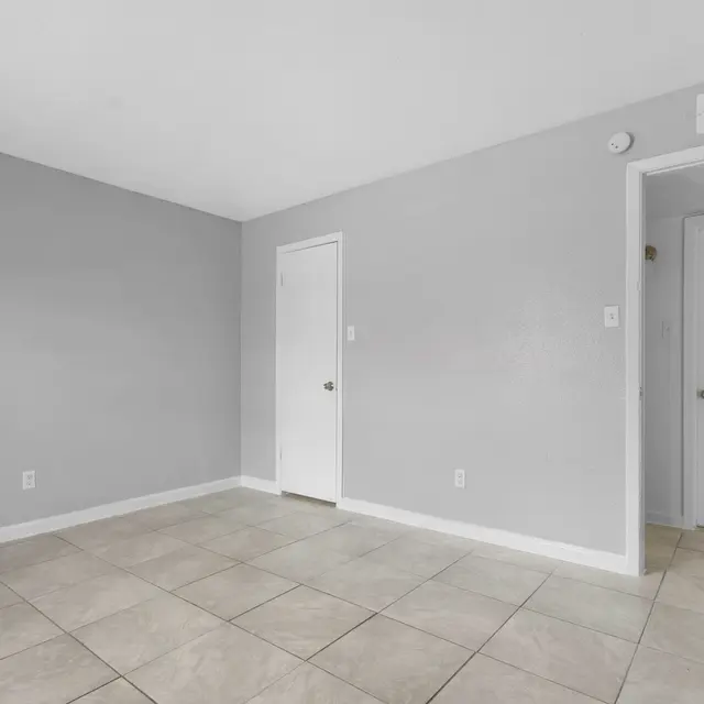 A spacious empty room with gray walls and tiled flooring, featuring a closed door on the left and an entrance to another room on the right.