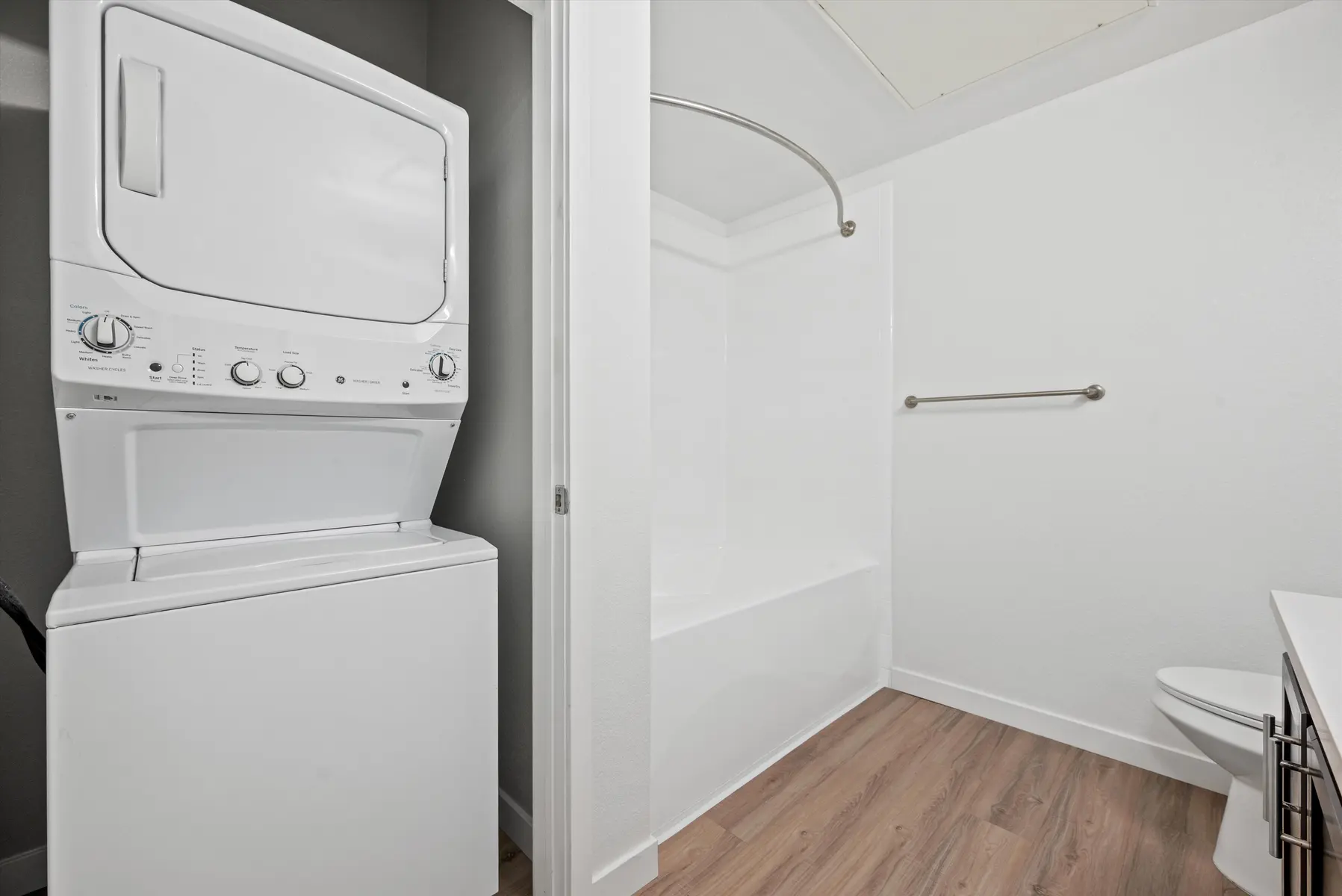 A modern bathroom featuring a stacked washer and dryer unit on the left, a shower tub combo with a curved curtain rod, and a toilet on the right side of the image.