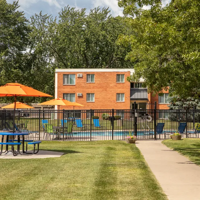 Regency Park - A view of a sunny poolside area featuring blue and green lounge chairs and umbrellas, surrounded by a black fence. In the foreground, a picnic table is placed on a grassy lawn, and a pathway leads towards the pool.